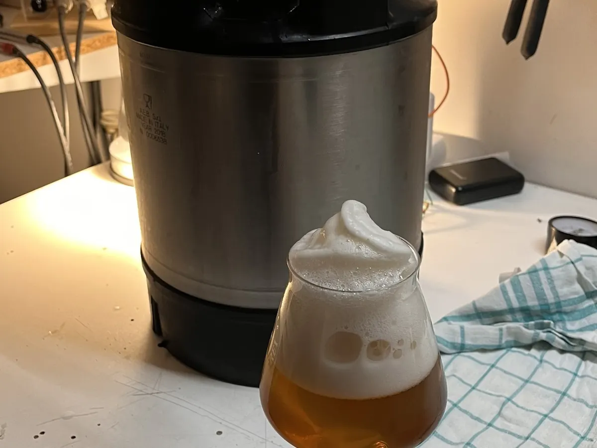 Glas Bier mit einer hohen, weichen Schaumkrone vor einem Keg
